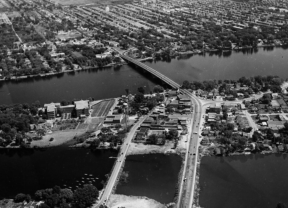 Vue aérienne en noir et blanc d’un pont qui enjambe une large rivière. Sur les deux rives, des quartiers habités.