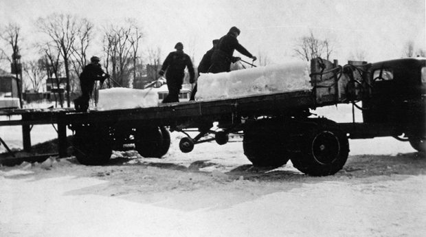 Photographie en noir et blanc d’une remorque sur laquelle quatre hommes chargent de gros blocs de glace.