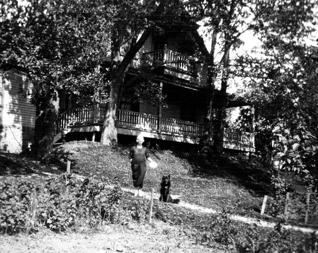 Photographie en noir et blanc d’une femme avec son chien sur un chemin devant une grande maison, en été.