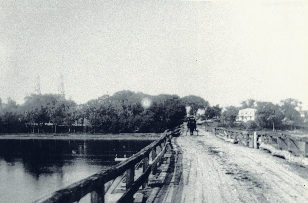 Photographie en noir et blanc du pont Bellefeuille.  Fait de bois, avec des balustrades sur les côtés, le pont ne suit pas une ligne droite. On voit une carriole à l’autre extrémité du pont.