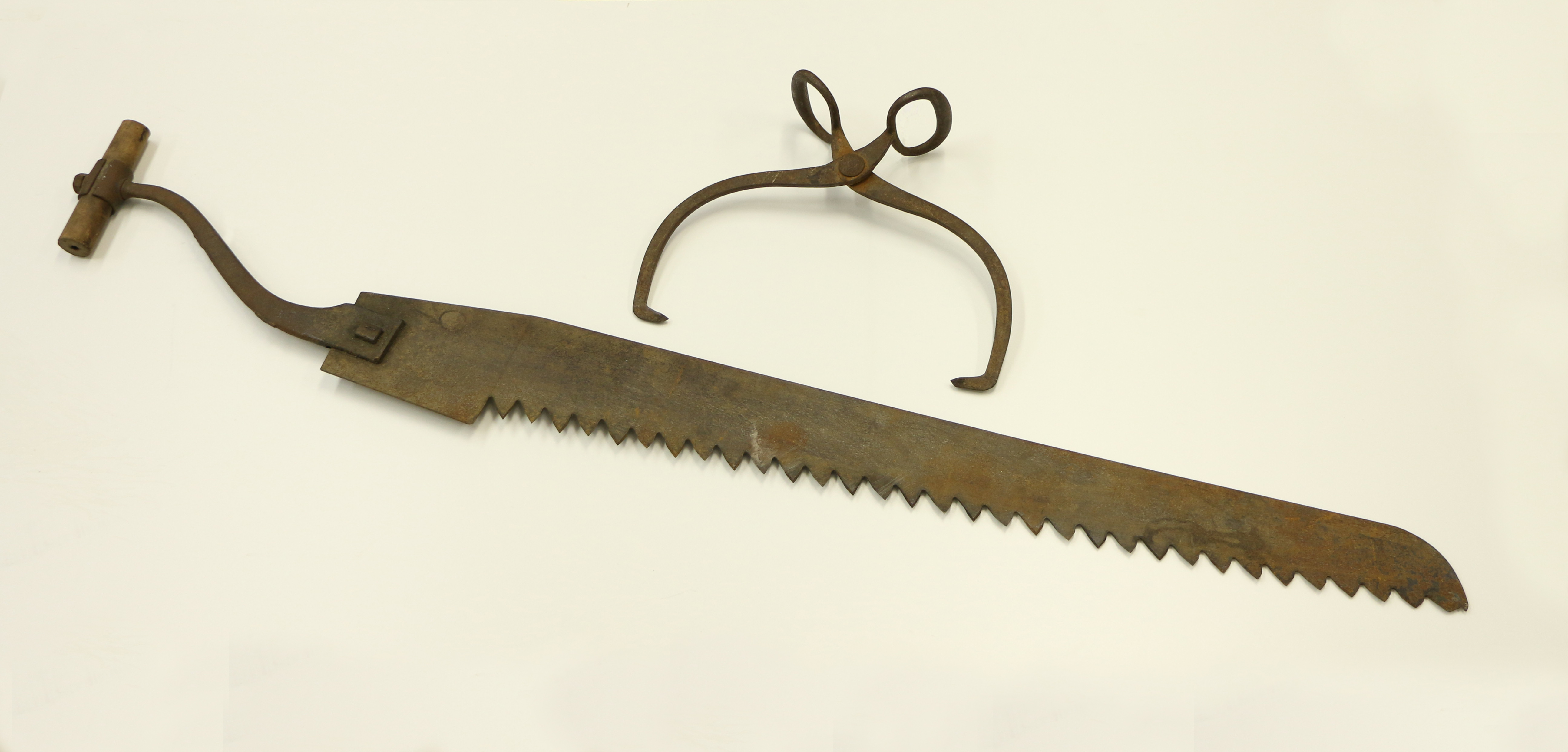 Photographie d’outils pour la coupe de glace. Sur un fond blanc, une pince en métal brun utilisée pour transporter les blocs et une scie en fer rouillé avec une poignée en bois.