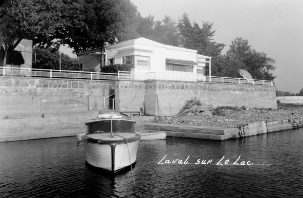 Photographie en noir et blanc d’une maison blanche et d’un petit bateau de plaisance amarré à un quai en pierres grises.