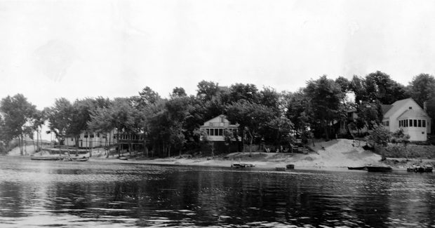 Photographie en noir et blanc montrant trois chalets en bois entourés d’arbres au bord d’une rivière.