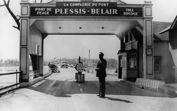 Photographie en noir et blanc de l’entrée du pont Plessis-Bélair. Une arche indique le nom du pont et la mention « pont à péage / toll bridge ». Un gardien est placé devant le poste de péage.