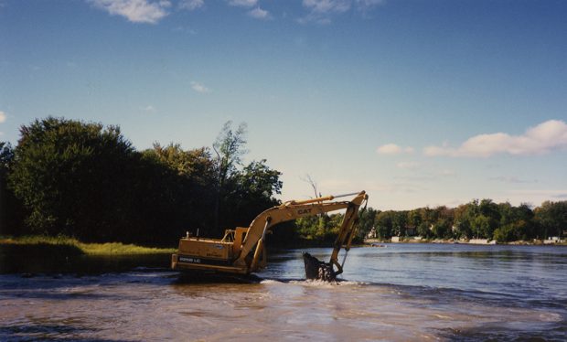 Photographie en couleur d’une pelle mécanique jaune travaillant dans la rivière des Prairies pour une opération de nettoyage lors d’une journée ensoleillée d’été.