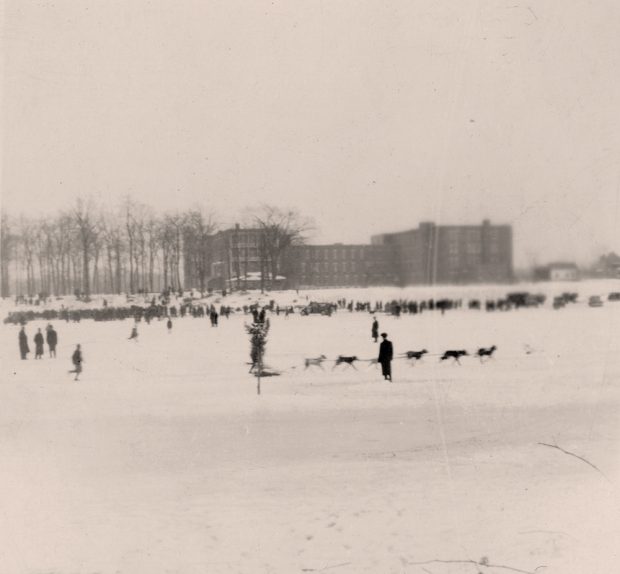 Photographie en noir et blanc d’une course de chiens sur la rivière des Prairies en hiver. Des centaines de personnes sont sur la rive enneigée de la rivière et six chiens, attelés les uns aux autres, tirent un traineau.