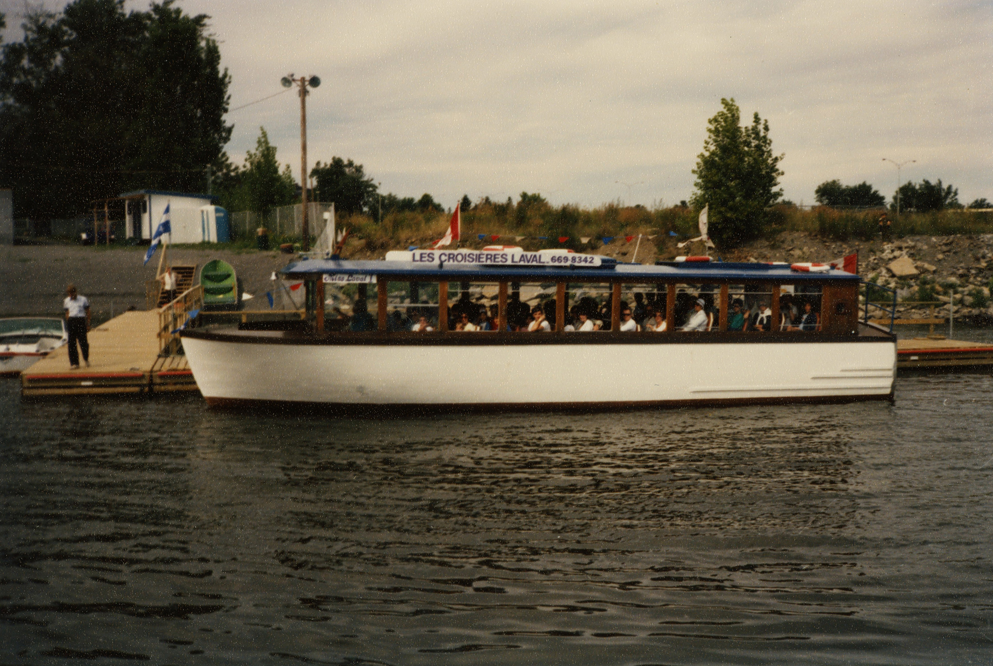 Photographie en couleur d’un bateau blanc accosté et rempli de plaisanciers prêts pour une croisière sur la rivière des Prairies.