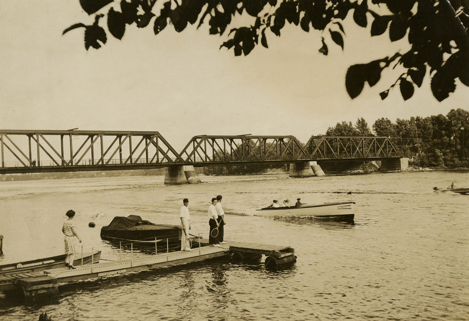 Photographie en sépia de gens sur un quai regardant des embarcations à moteur sur la rivière. En arrière-plan, un pont pour les trains enjambe la rivière.