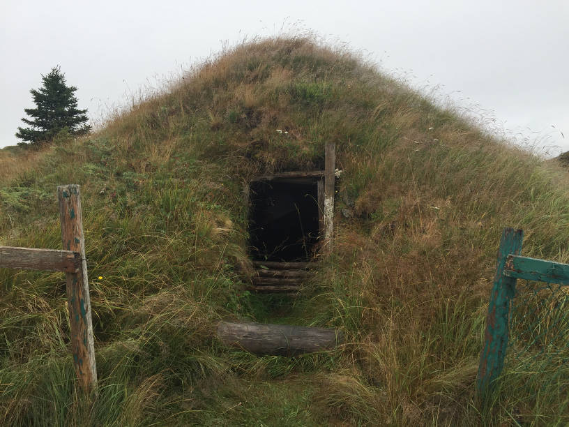 Fosse encadrée de bois au centre d’une grande butte herbeuse avec, à l’avant, une marche faite d’un rondin.