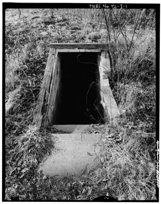 Au milieu d’un champ d’herbes hautes, ouverture rectangulaire encadrée de bois menant au fond d’une fosse aux murs de pierre à l’aide d’un escalier en béton.