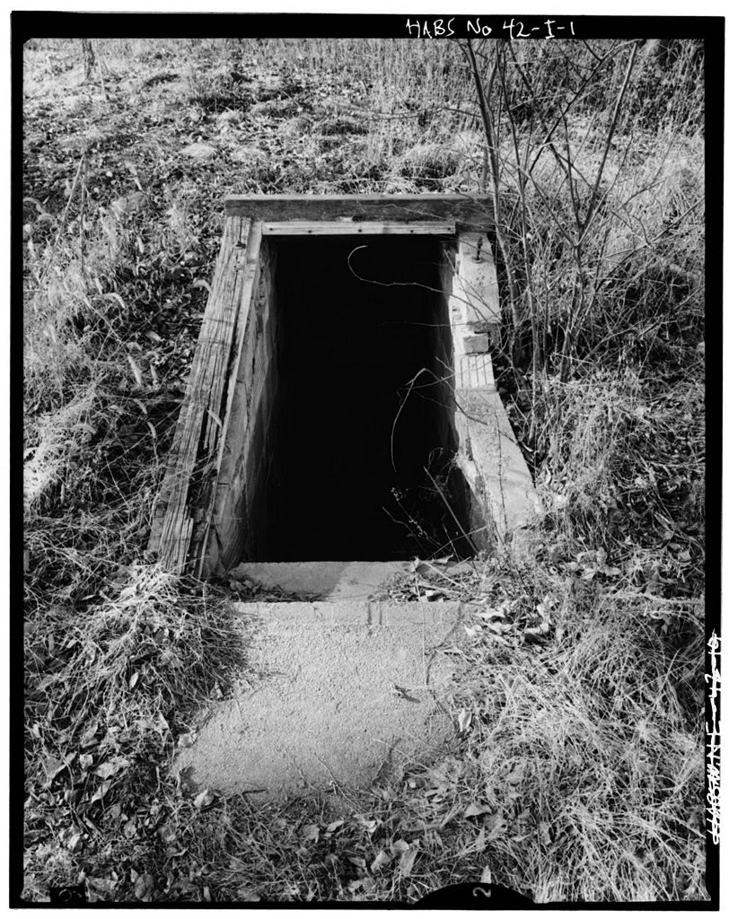 Au milieu d’un champ d’herbes hautes, ouverture rectangulaire encadrée de bois menant au fond d’une fosse aux murs de pierre à l’aide d’un escalier en béton.