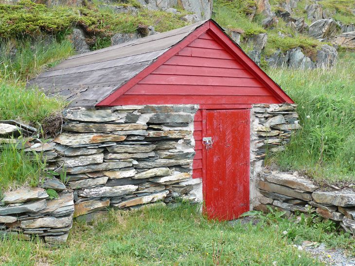 Cave à légumes en pierres empilées à flanc de colline, avec porte en bois rouge et remise à toit rouge dans un champ herbeux.