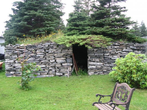 Extérieur d’une cave à légumes en pierres empilées avec un toit en terre recouvert d’herbe et d’arbustes.