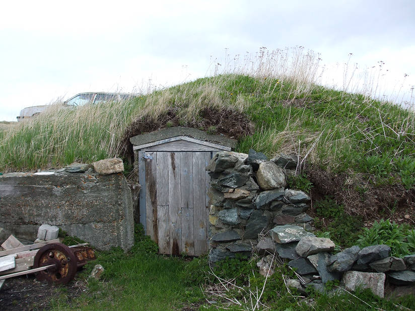 Extérieur d’une cave à légumes à flanc de colline avec un mur en béton (à gauche), une porte d’entrée en bois (au centre) et un tas de pierres empilées (à droite).