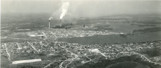 Photo noir et blanc de deux agglomérations, séparées par un terrain vague, construite autour d’un lac. À l’arrière-plan, la fonderie avec trois chevalements et deux cheminées qui fument.