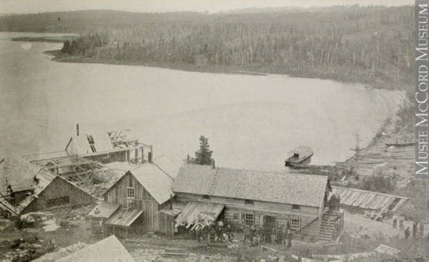 Photographie en noir et blanc d’une quinzaine de personnes devant les quatre bâtiments en planche de la mine Wright situés sur le bord du lac Témiscamingue.