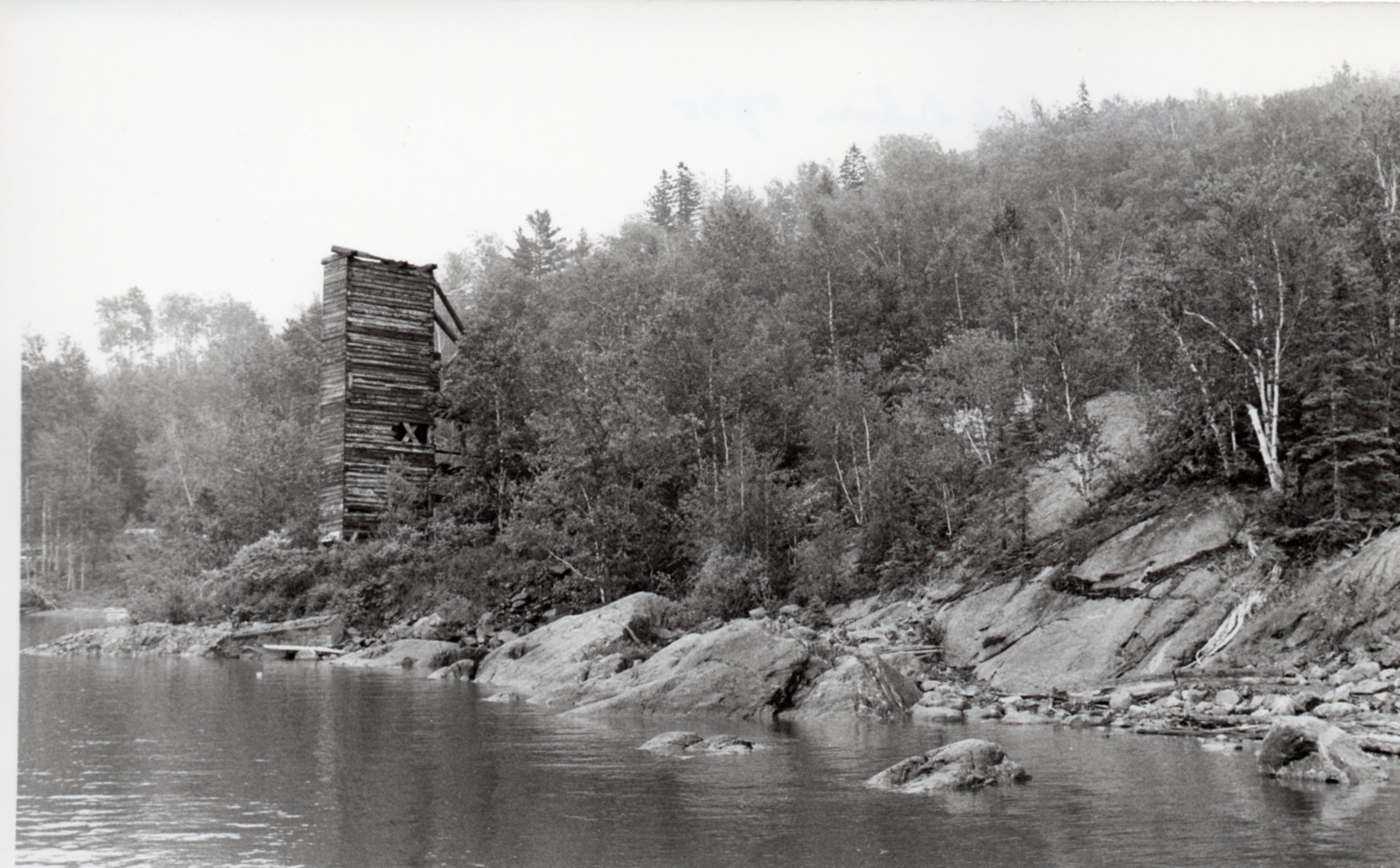 Photographie en noir et blanc de la rive, recouverte de rocher et d’arbres, du lac Témiscamingue avec une haute structure de planche en forme de tour carrée.