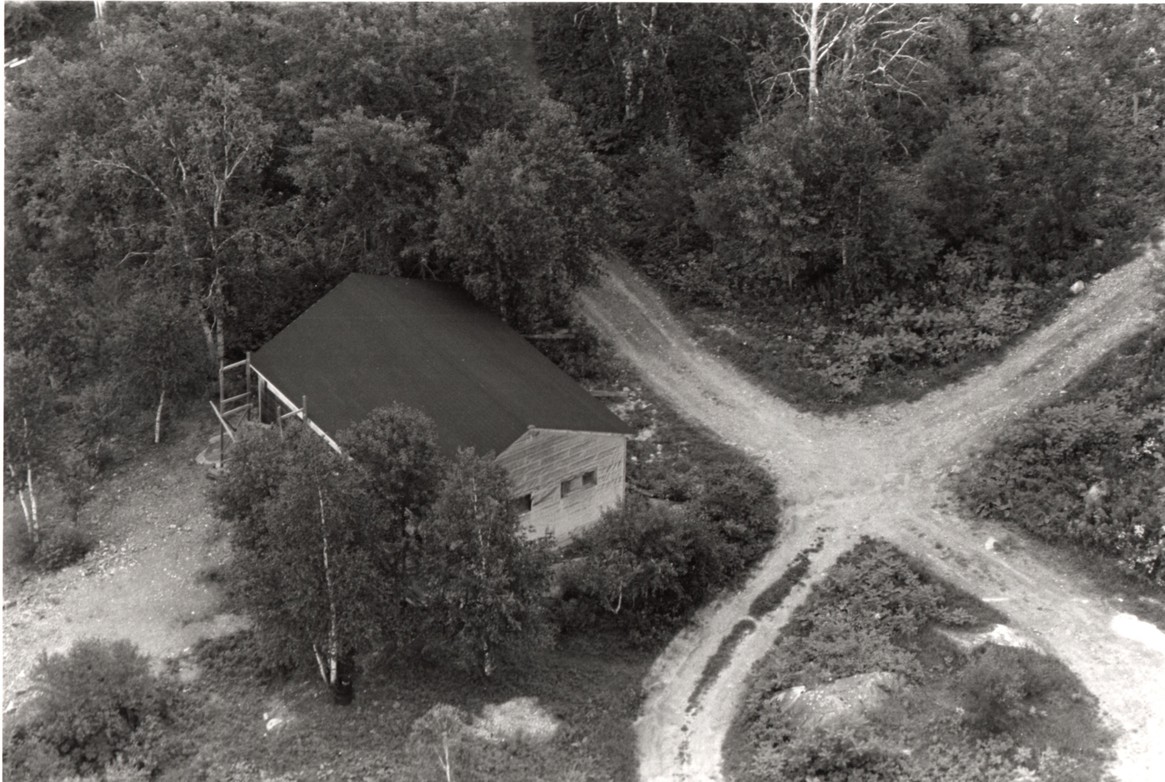 Photographie noir et blanc, vue aérienne d’un bâtiment en planche entouré d’arbres situé à la croisée de deux chemins de graviers.