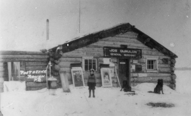 Photographie en noir et blanc d’une cabane en bois rond avec l’inscription Jos Dumulon General Merchant. En avant-plan, un jeune garçon ainsi que deux chiens. 