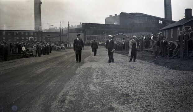 Photographie en noir et blanc d’un chemin avec des gens rassemblé sur les côtés et cinq policiers, dont deux en uniforme, au centre. La fonderie Horne est en arrière-plan.