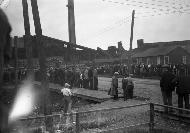 Photographie en noir et blanc de gens rassemblés sur deux trottoirs parallèle devant la fonderie Horne qui est en arrière-plan.