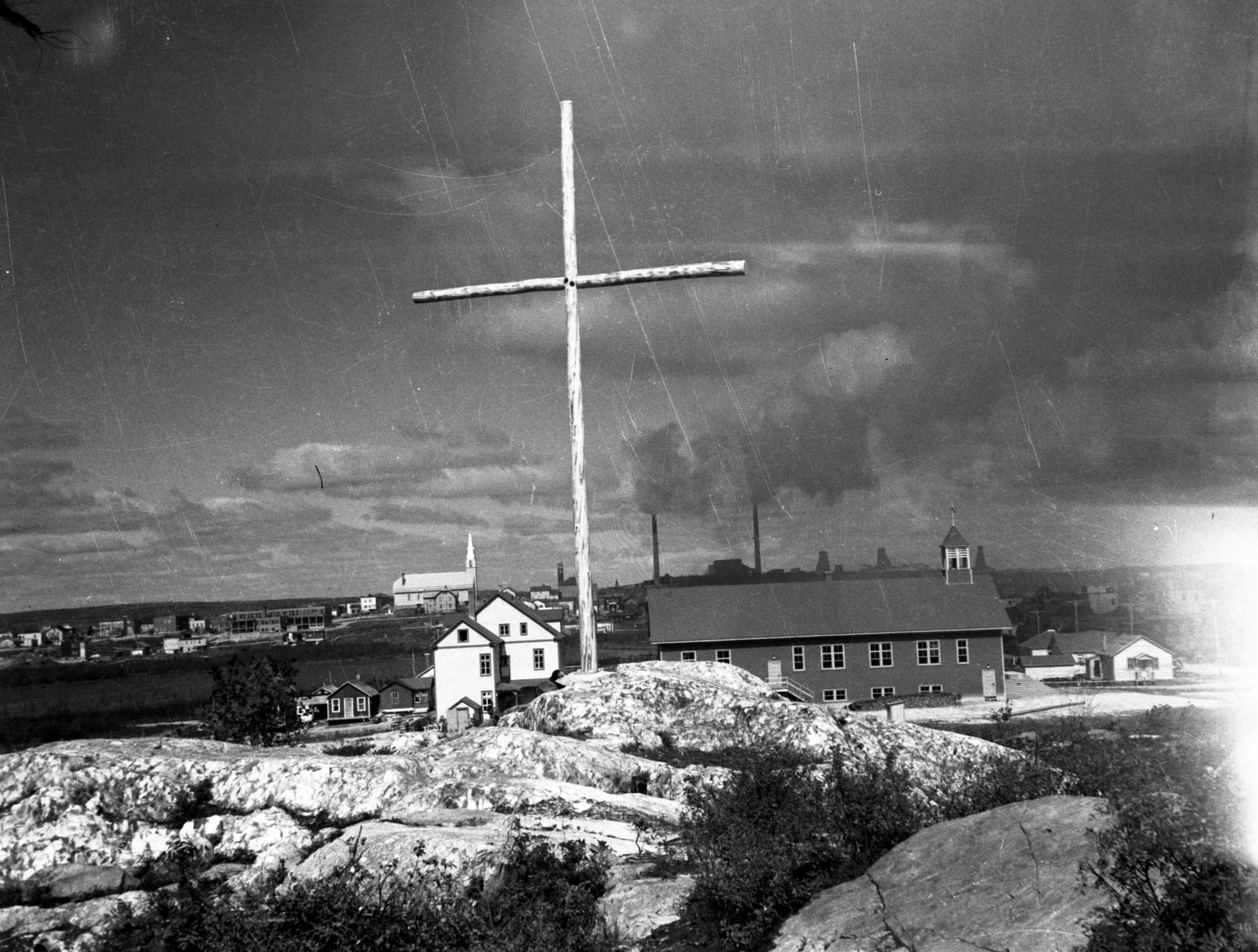 Photographie en noir et blanc  de rochers, d’une croix, d’une église et d’un presbytère. En arrière-plan, à gauche, la ville de Rouyn, et à droite, les installations de la mine Horne.