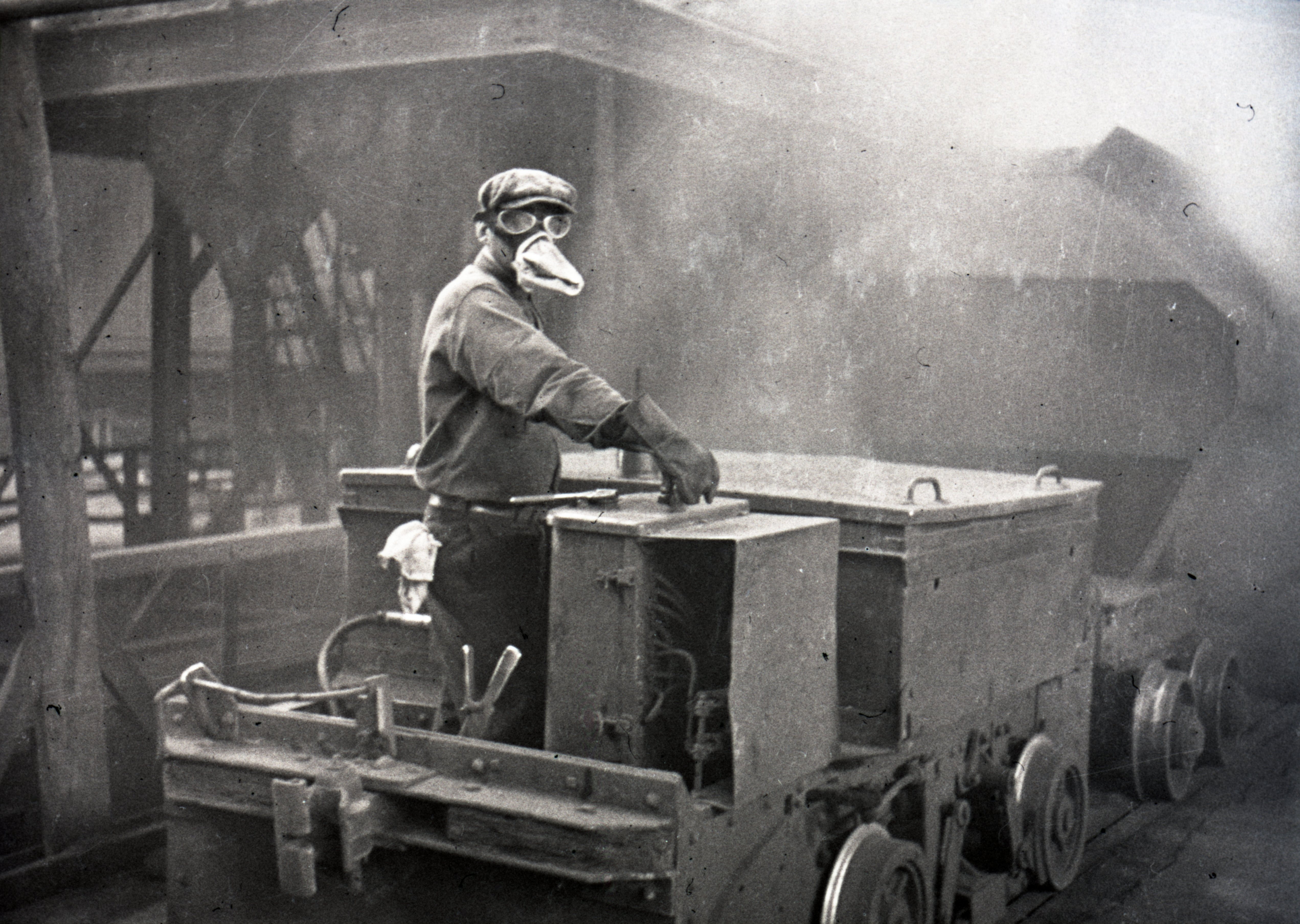 Photographie en noir et blanc d'un ouvrier portant un masque protecteur et des lunettes conduit un wagon à l’intérieur de la fonderie de la mine Horne. La présence de gaz rend la photo plutôt floue, mais on perçoit des structures en acier.