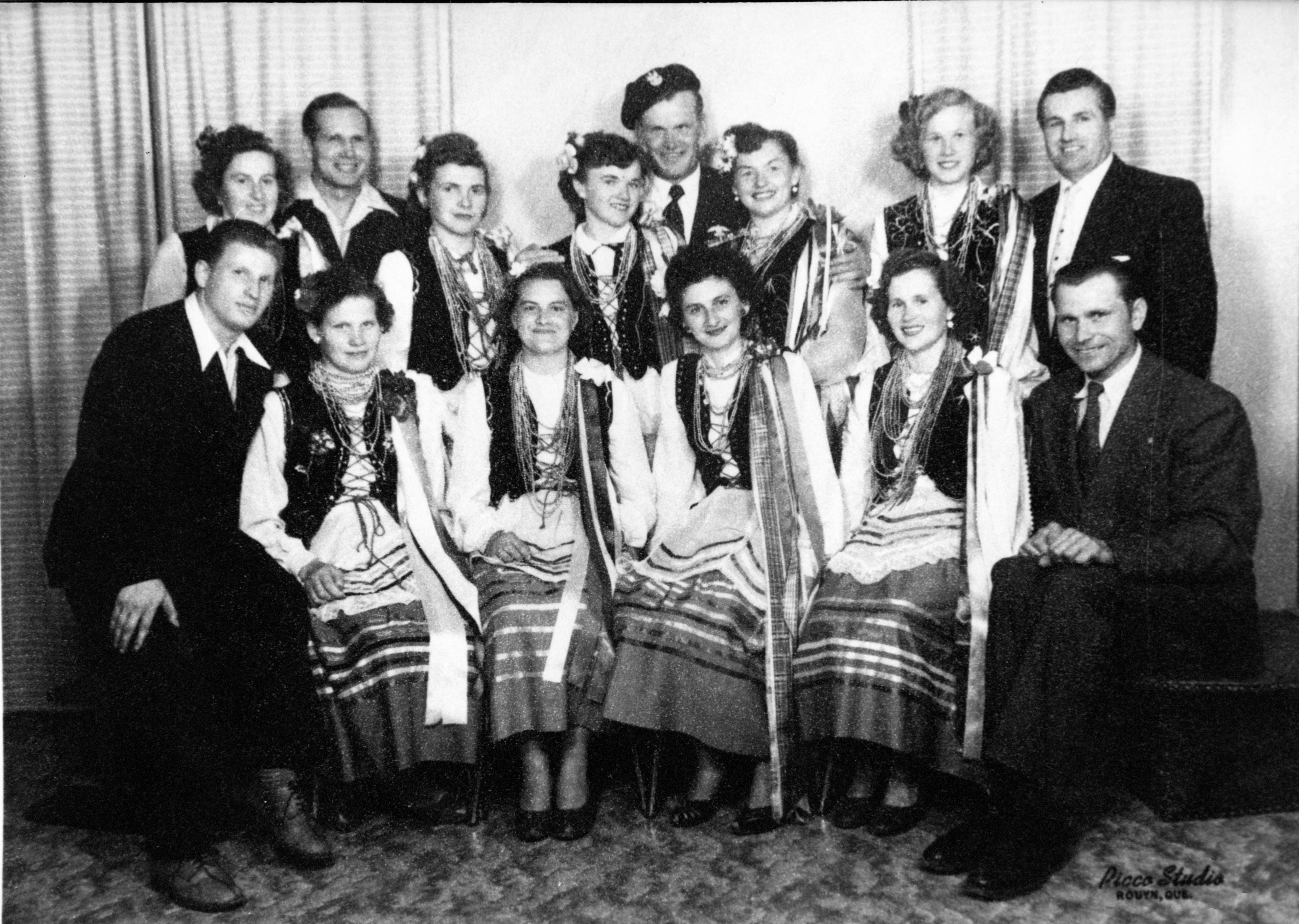 Photographie en noir et blanc de quatorze personnes, dont neuf femmes en habit traditionnel ukrainien et cinq hommes en habit chic.