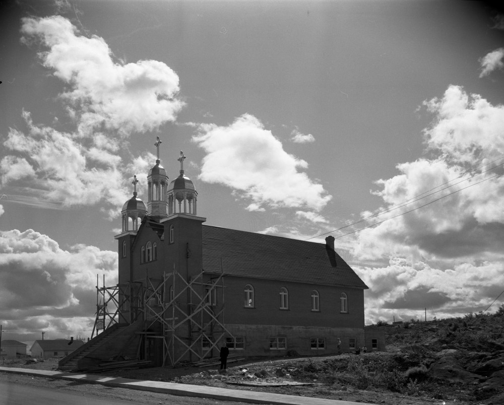 Photographie en noir et blanc d’une église, avec trois clochers et trois croix, dont la façade est entourée d’un échafaud. À l’avant, un escalier se rend aux portes principales.