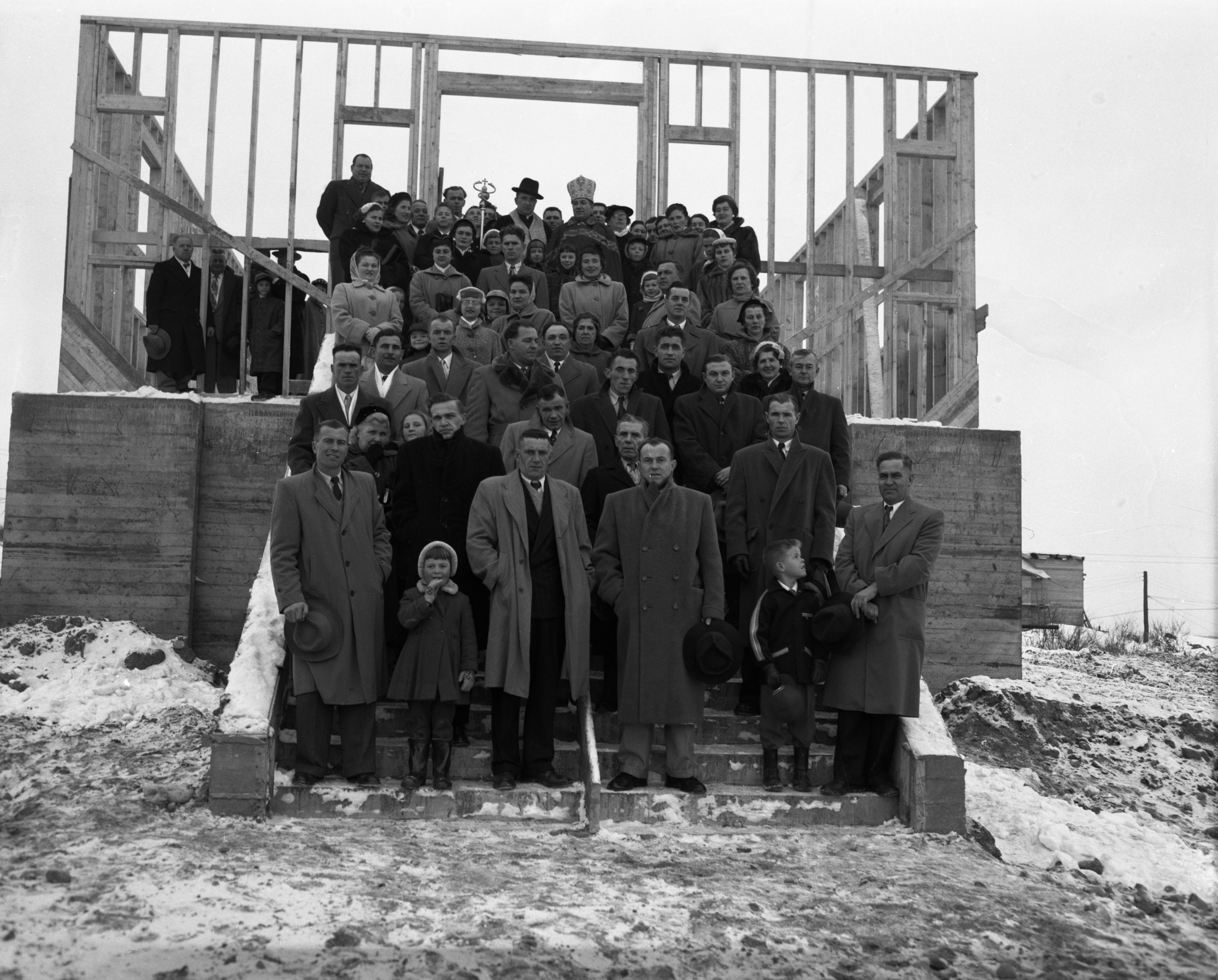 Photographie en noir et blanc d’environ soixante-dix personnes dans des escaliers devant une structure de bois. Dont deux prêtres catholiques, un romain et un ukrainien. 