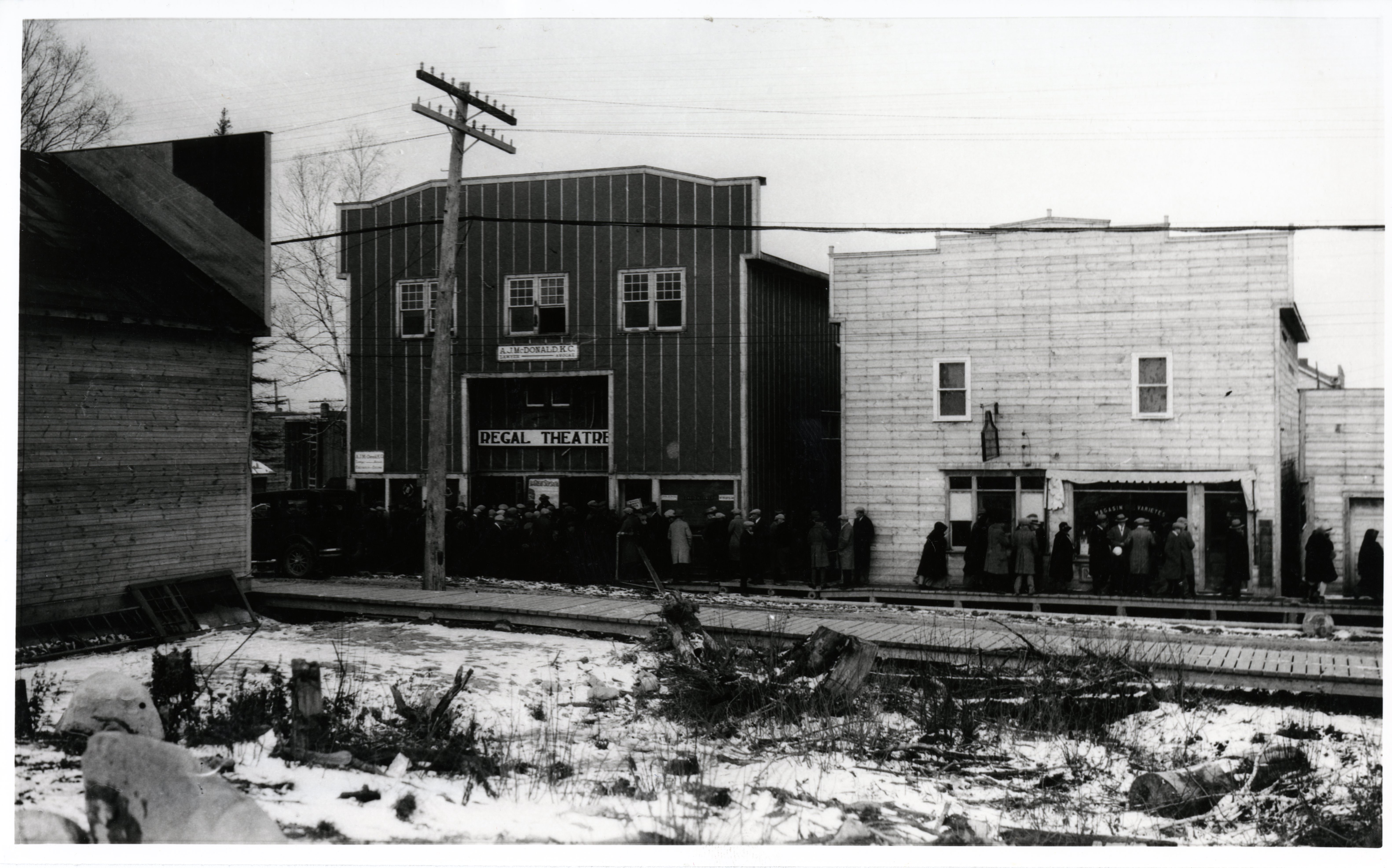 Photographie en noir et blanc de deux bâtiments avec des façades Boomtowns, dont un qui a une affiche Regal Theatre. Une foule attend sur le trottoir de bois devant l’édifice.