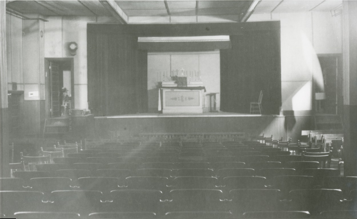 Photographie noir et blanc d’une salle de théâtre, avec des chaises fixes et d’autres mobiles, dont la scène est occupée par un autel sur lequel est disposé un tabernacle.