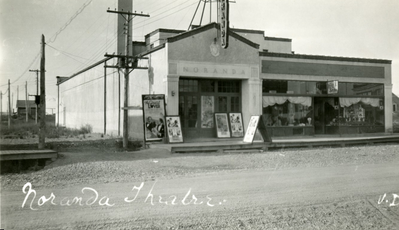 Photographie en noir et blanc d’un bâtiment, avec le nom Noranda inscrit sur la façade, et plusieurs affiches de films installées sur le trottoir de bois. 