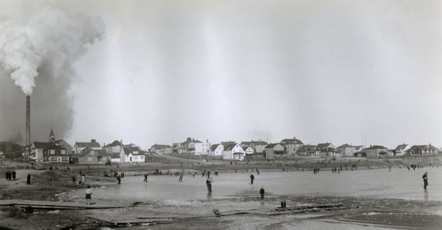 Photographie en noir et blanc d’un lac gelé, mais sans neige, sur lequel une quarantaine de personne patine. En arrière-plan, la ville de Noranda et une cheminée de la fonderie.