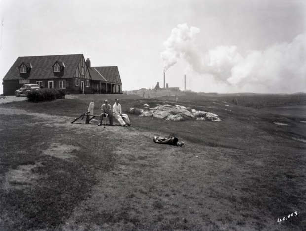 Photographie en noir et blanc de deux hommes assis devant le pavillon du club de golf de Noranda avec, en arrière-plan, la mine Horne.