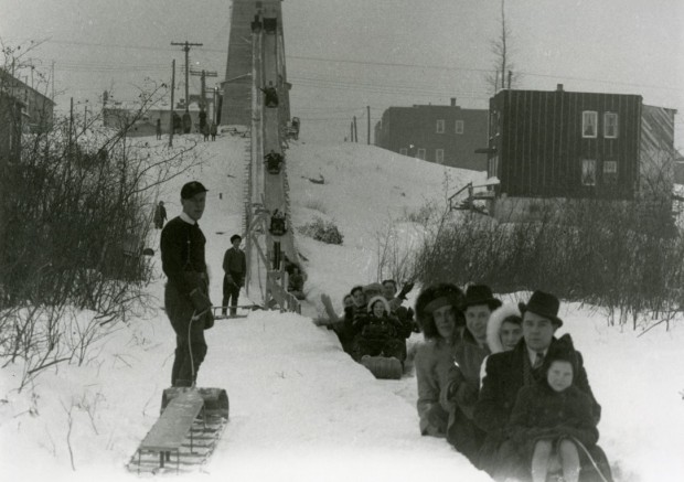 Photographie en noir et blanc de plusieurs personnes sur des traines sauvages qui glissent à partir d’une structure de bois installée sur une colline. 