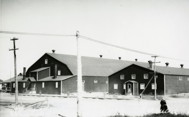 Photographie en noir et blanc de deux bâtiments, avec des toits de tôle et très peu de fenêtres. Sur le sol, une fine couche de neige.