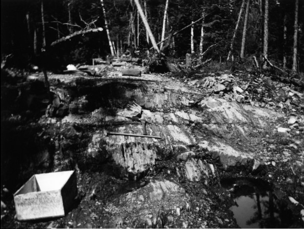 Photo en noir et blanc d’un sol rocheux, ainsi que de deux pioches et de deux boites de bois, avec la forêt en arrière-plan. 