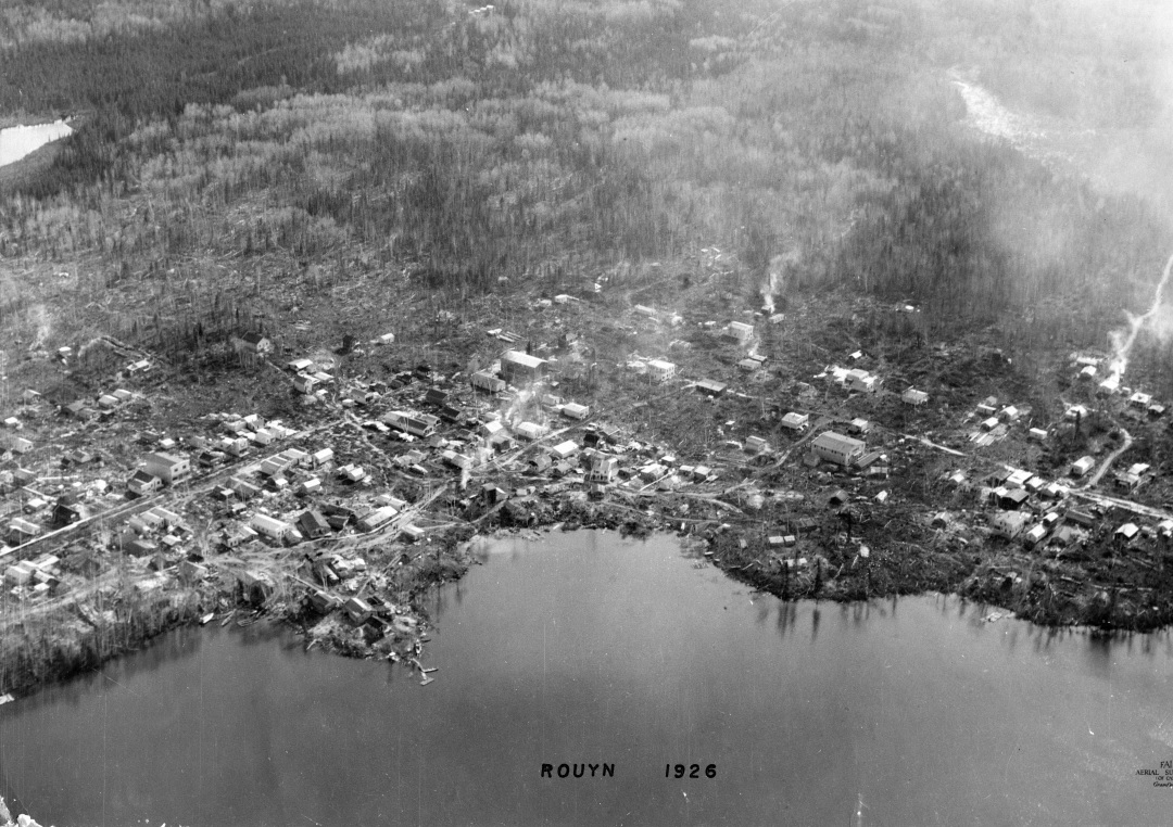 Photographie en noir et blanc d’une centaine de cabanes en bois rond installées de façon anarchique sur le bord du lac Osisko.