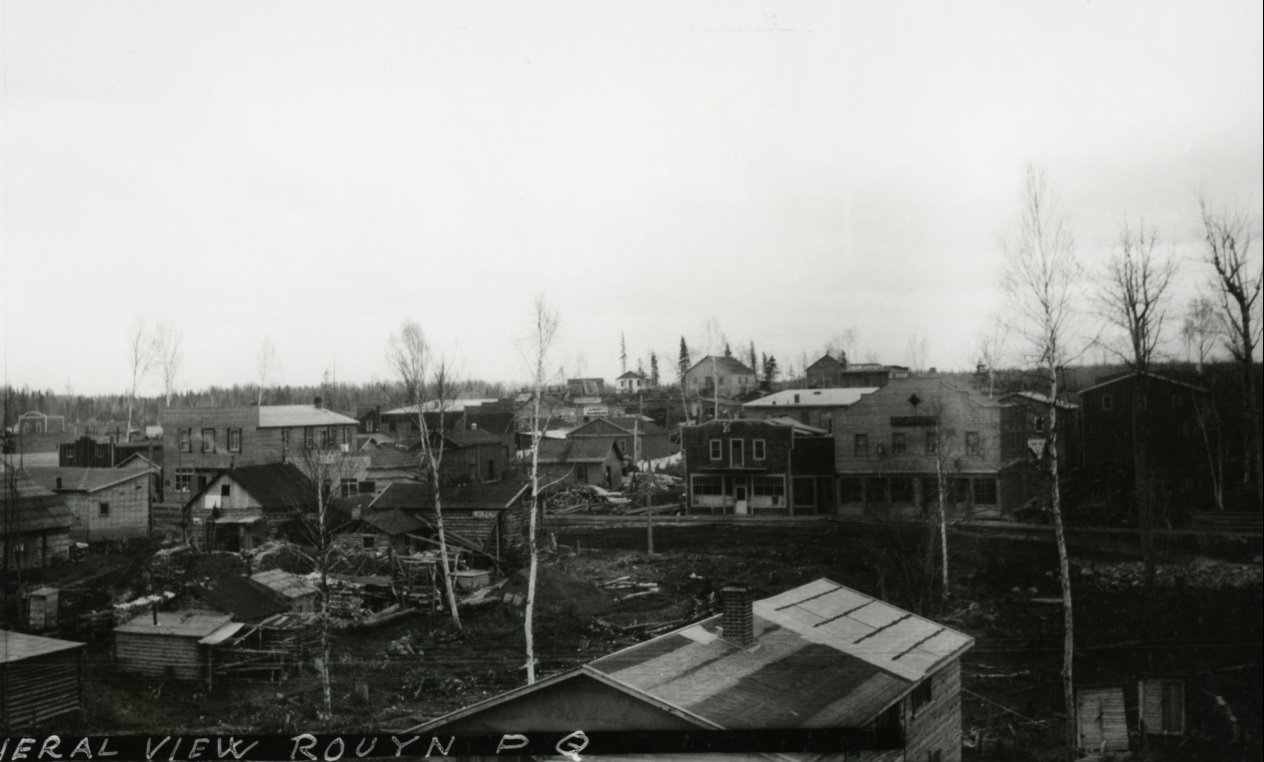 Photographie en noir et blanc de nombreuses cabanes, en rondins ou en planches, ainsi que d’un chemin de terre à peine défriché. Plusieurs arbres se dressent entre les bâtiments.