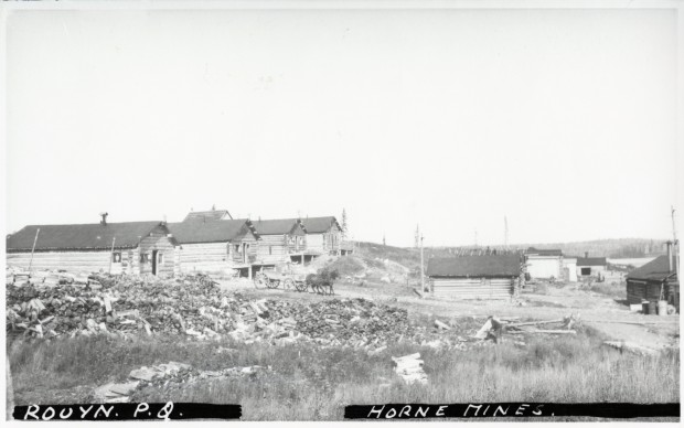 Photographie en noir et blanc de plusieurs cabanes en bois rond, d’une charrette tirée par deux chevaux et de nombreux amoncellements de bûches.