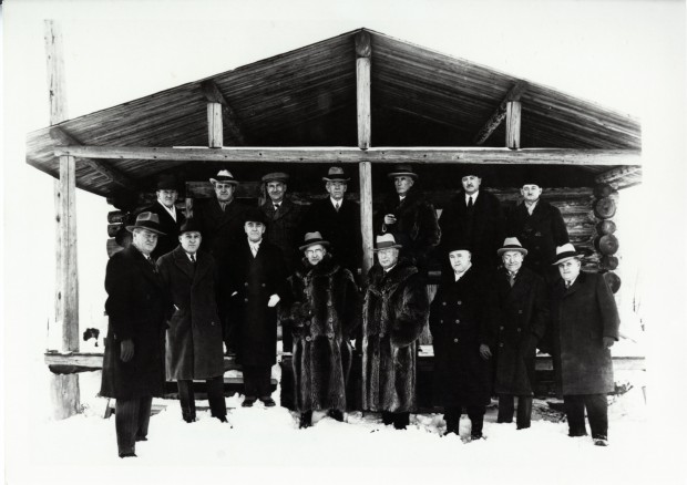 Photographie noir et blanc de quinze hommes en habit chic, dont trois avec des manteaux de fourrure, devant une cabane en bois rond.
