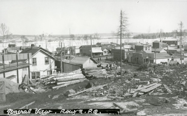 Photographie en noir et blanc de bâtiments rudimentaires construits au sud-ouest du lac Osisko. En avant-plan, des roches, des bûches, des planches et des débris.