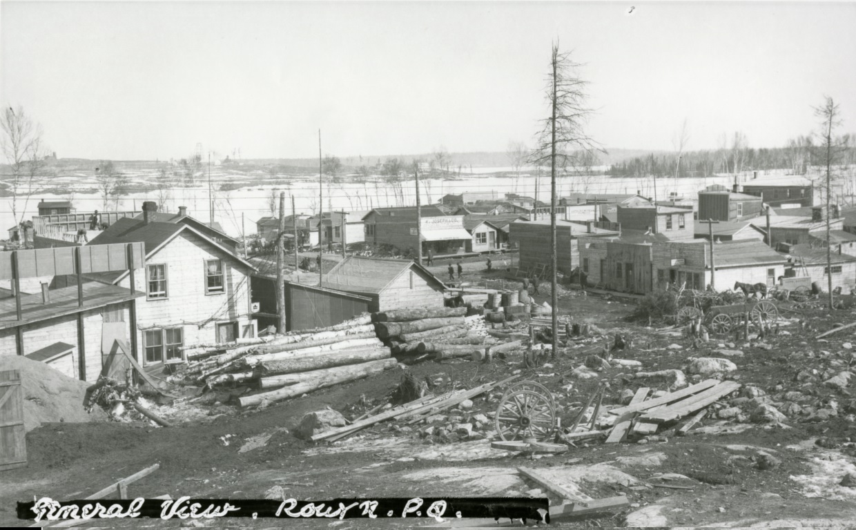 Photographie en noir et blanc de bâtiments rudimentaires construits au sud-ouest du lac Osisko. En avant-plan, des roches, des bûches, des planches et des débris.