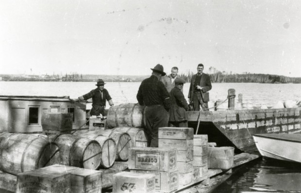 Photographie en noir et blanc de cinq hommes, sur un quai, entrain de décharger des boites et des tonneaux de bois.