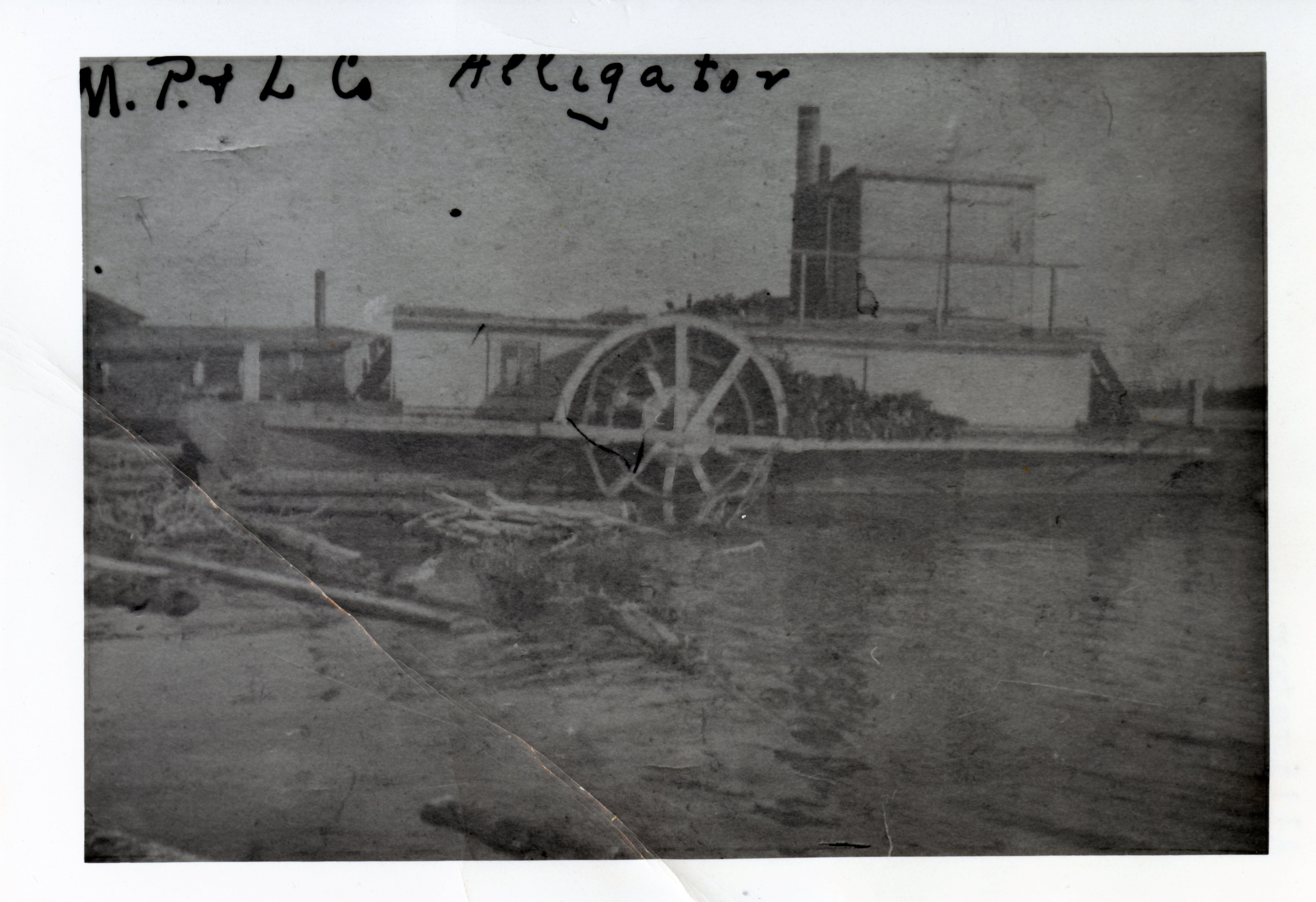 Photographie en noir et blanc, de mauvaise qualité, d’un bateau à vapeur qui est accosté. Sur la rive, un bâtiment et des billots flottent sur l’eau.