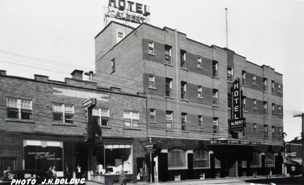 Photographie en noir et blanc d’un bâtiment à quatre étages avec plusieurs enseignes, dont une sur le toit, qui indique Hotel Albert. À gauche, un édifice à deux étages.