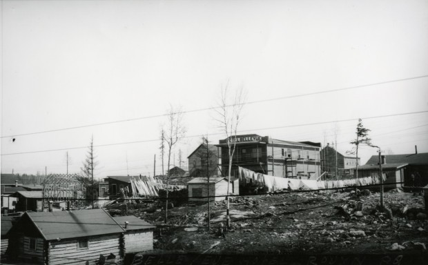 Photographie en noir et blanc de plusieurs bâtiments rudimentaires, en planche ou en bois rond, dont l’un avec l’inscription Hôtel Bellevue sur la façade de style Boomtowns.