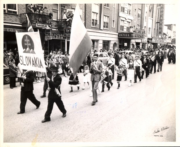 Photographie en noir et blanc d’environ vingt-cinq personnes qui défilent, devant une foule sur le trottoir de la rue Principale, avec une affiche portant l’inscription Slovakia surmonté du blason national et d’un drapeau de la Slovaquie.