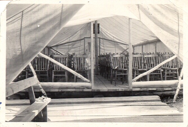 Photographie en noir et blanc de l’intérieur d’une tente  dans laquelle des chaises, disposées en rangées, sont installées sur un plancher de bois.